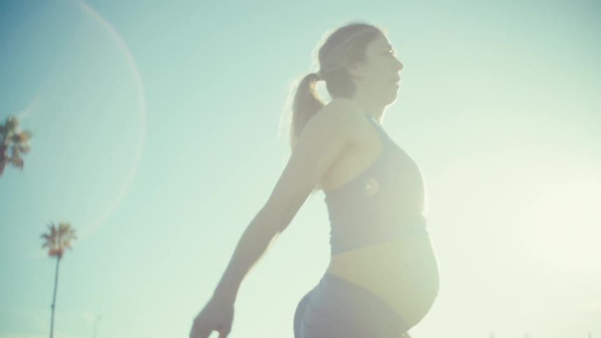 A young pregnant woman stretching her feet outdoors under the sun with her baby bump visible