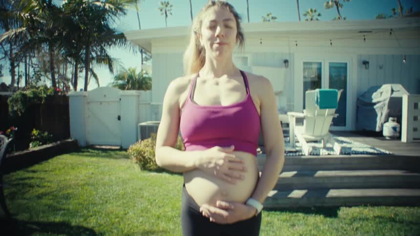 A pregnant woman standing on a yoga mat in her backyard holding her baby bump while meditating at midday