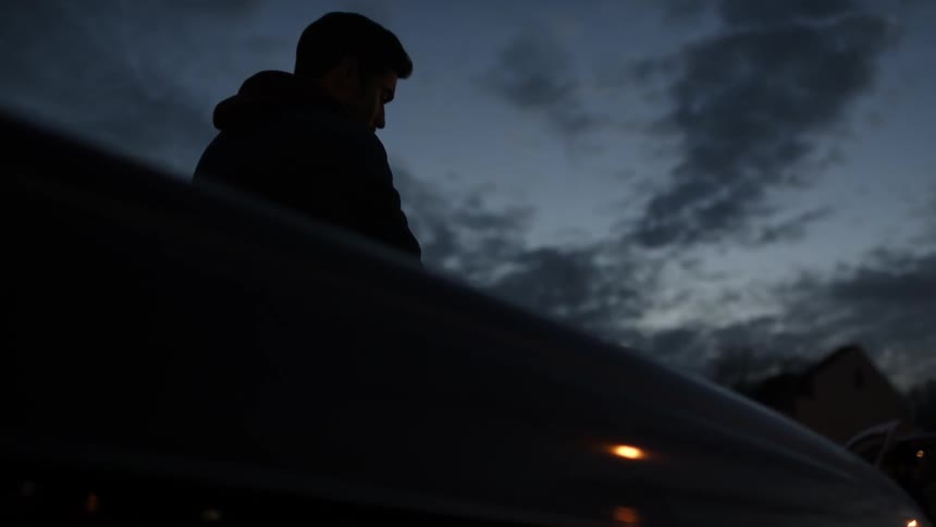 A lone man leans against a vehicle parked in a neighborhood at dusk