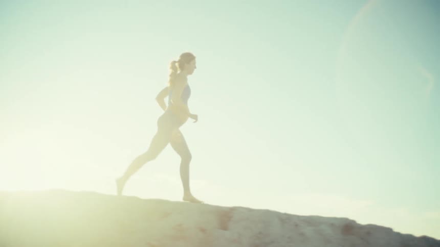 A pregnant woman in athletic wear runs across the crest of a sandy hill against a bright sundrenched sky as she moves from left to right the camera captures her silhouette and steady pace in the hazy daylight