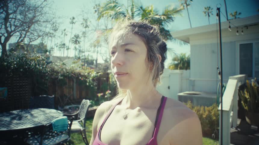 A pregnant woman holding her belly while standing in a sunny backyard with a house and palm trees behind