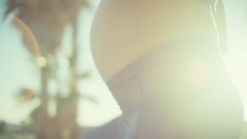 A pregnant woman jogging outdoors under the sun wearing blue tight pants with her baby bump visible