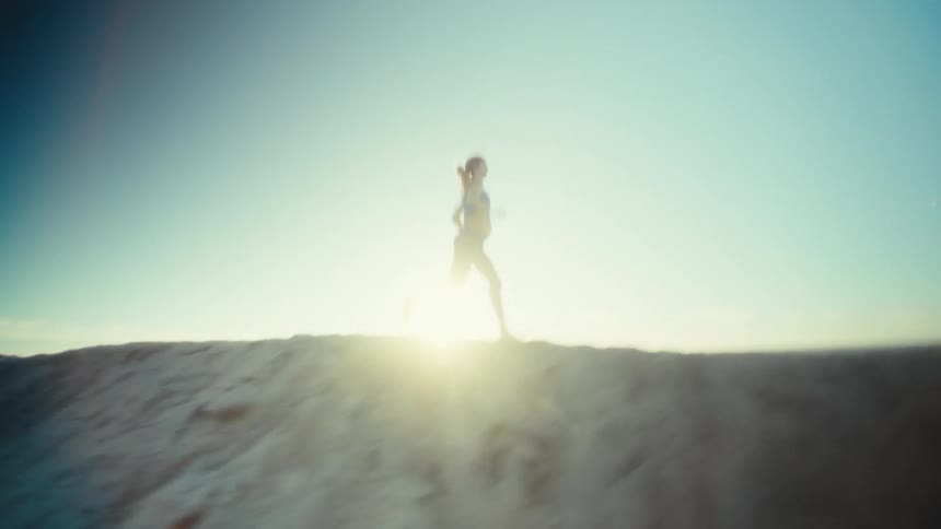 A lone runner moves along the crest of a sandy ridge silhouetted against a vast blue sky as sunlight flares behind each determined stride