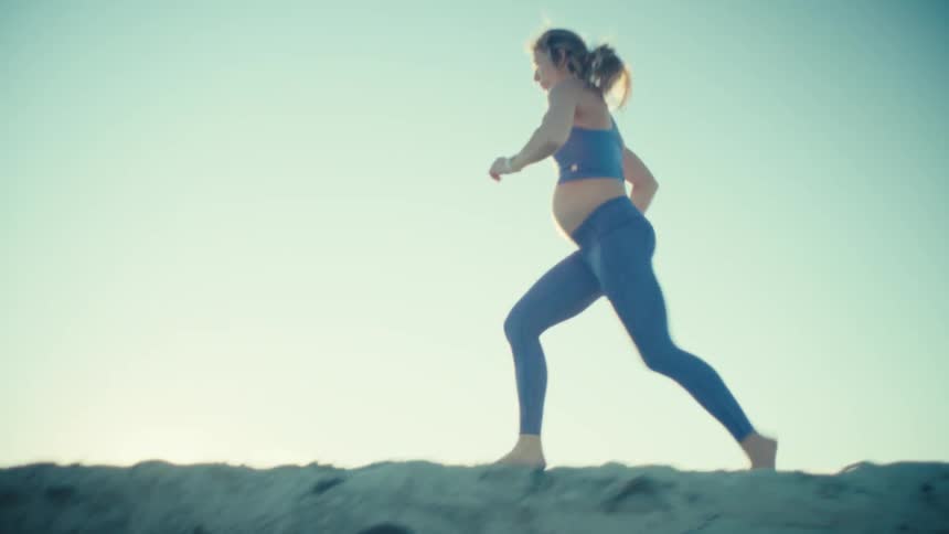 A pregnant woman wearing a blue sports bra and leggings runs along a sandy beach during a bright sunset