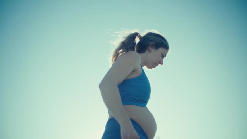 A pregnant woman in blue athletic wear performs leg stretches outdoors under a bright clear skythe sun creates a lens flare as she balances on one leg preparing for a workout