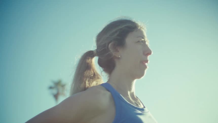 A pregnant woman in a blue sports top stretches her leg while standing in a sunny outdoor areathe camera circles around her to show her face and baby bump against a backdrop of palm trees and a clear blue sky