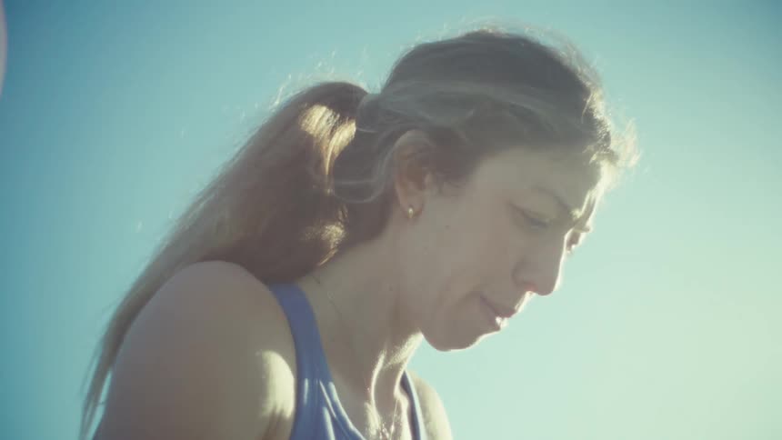 A pregnant woman wearing a blue sports top and leggings stretches her legs while standing in a grassy park with palm trees and a parking lot visible in the background under a clear sky