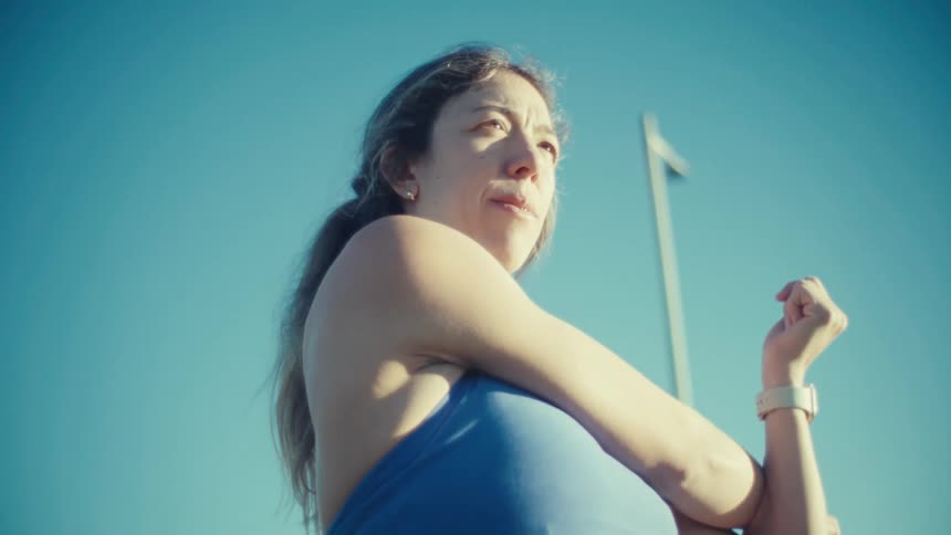 A pregnant woman in athletic wear performs arm stretches during an outdoor workout on a sunny day