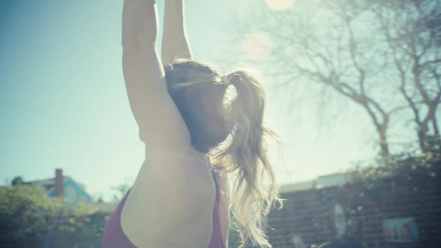 A pregnant woman in a pink sportsbra performs yoga stretches in a sunny backyard with her arms raised toward the sky while trees and a white house stand in the background