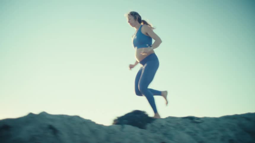 A young pregnant woman with a visible baby bump running barefoot on a sand hill on a sunny day