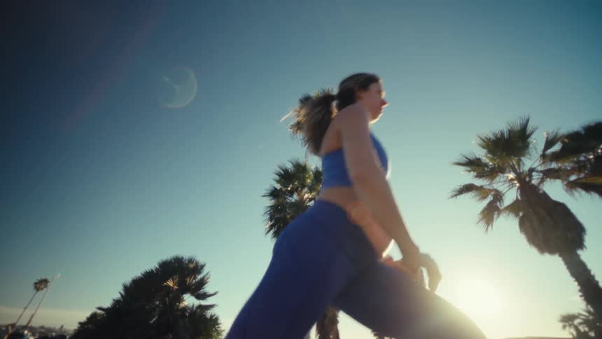 A view from a low angle shows a pregnant woman wearing a blue sports bra and blue leggings as she performs lunges in an outdoor area with palm trees under a clear blue sky