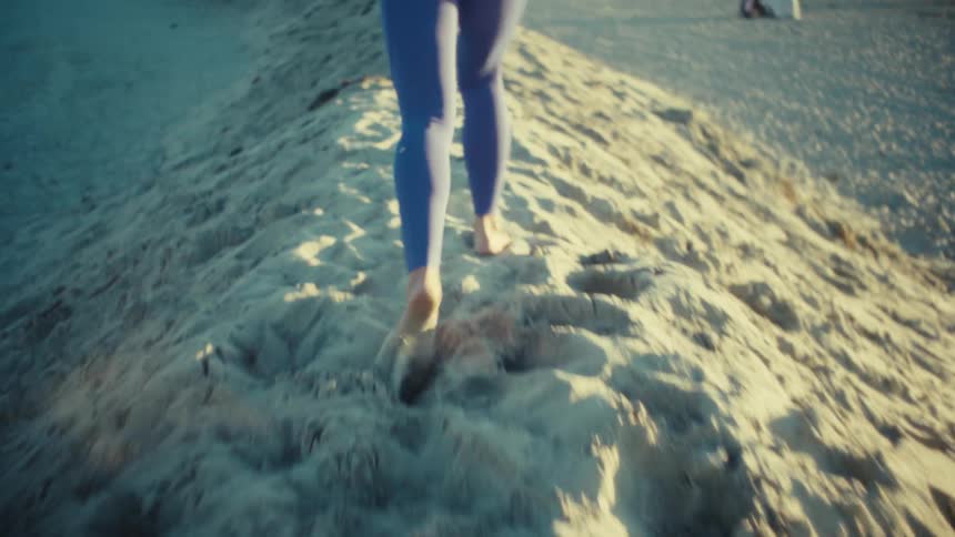 A view from behind shows a woman wearing blue athletic clothing running barefoot up a tall sand dune at a beach during a clear day with a coastal town visible in the distance