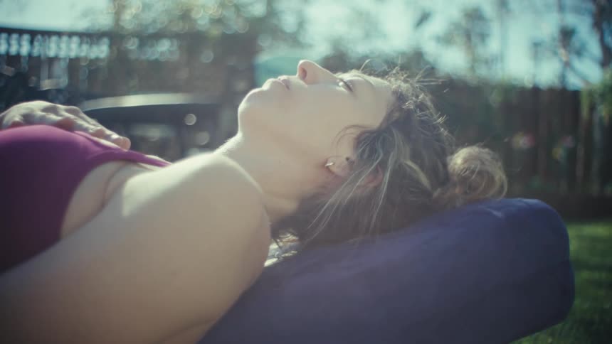 A woman lies back on a yoga bolster in a sunlit outdoor setting resting one hand on her stomach and the other on her chestshe keeps her eyes closed in a relaxed state breathing deeply while soaking in the sunlight