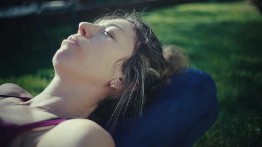 A woman wearing a maroon sports bra lies on a yoga mat in a sunlit backyard with her eyes closed while resting her head on a blue bolster
