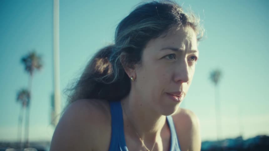 A pregnant woman wearing a blue sports bra and leggings exercises outdoors under a clear sky and the shot moves from her stomach to a close up of her face while cars and palm trees