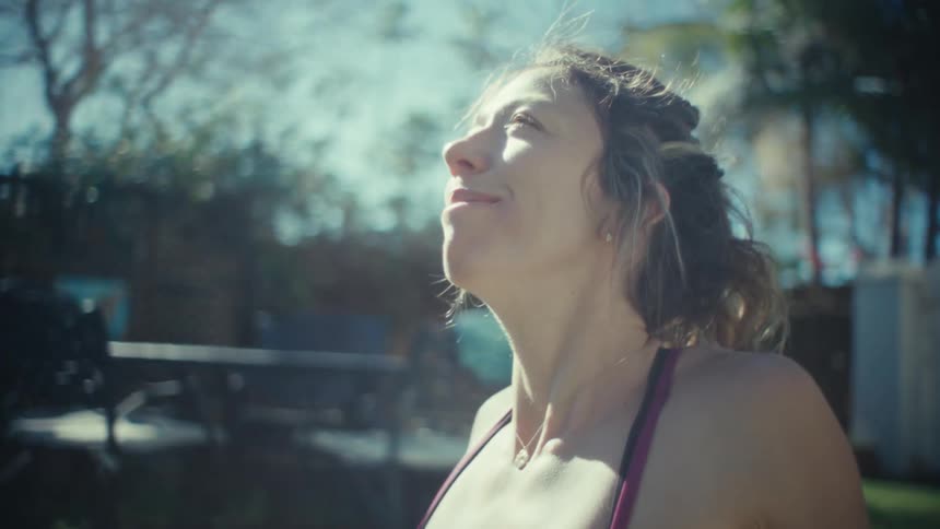 A woman wearing a maroon sports bra and black leggings sits on a yoga mat in a sunny backyard and smiles while raising her arms above her head and bringing her hands together