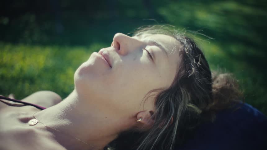 A woman lies peacefully on a yoga bolster in the grass with her eyes closedthe camera slowly zooms in on her face as she basks in the warm natural sunlight