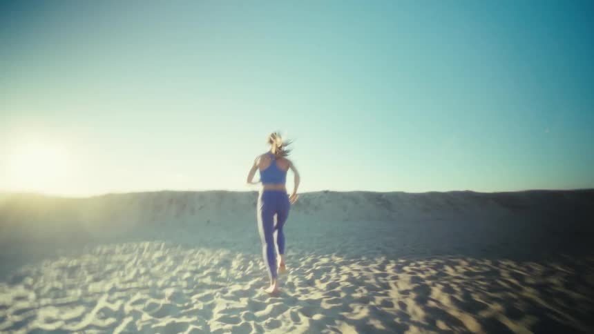 A woman in blue athletic gear runs across a sandy beach toward the bright glow of the setting sunthe camera follows from behind as she heads toward a sand dune capturing the energetic movement of her run against the wide coastal landscape