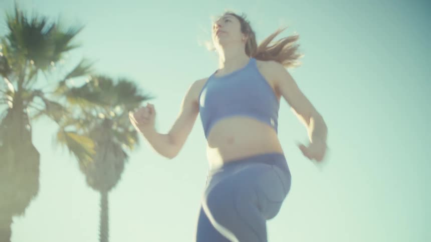 Videoa view from the front shows a pregnant woman wearing a blue sports top and leggings as she performs high knee exercises while running in place on a sandy beach during a bright day