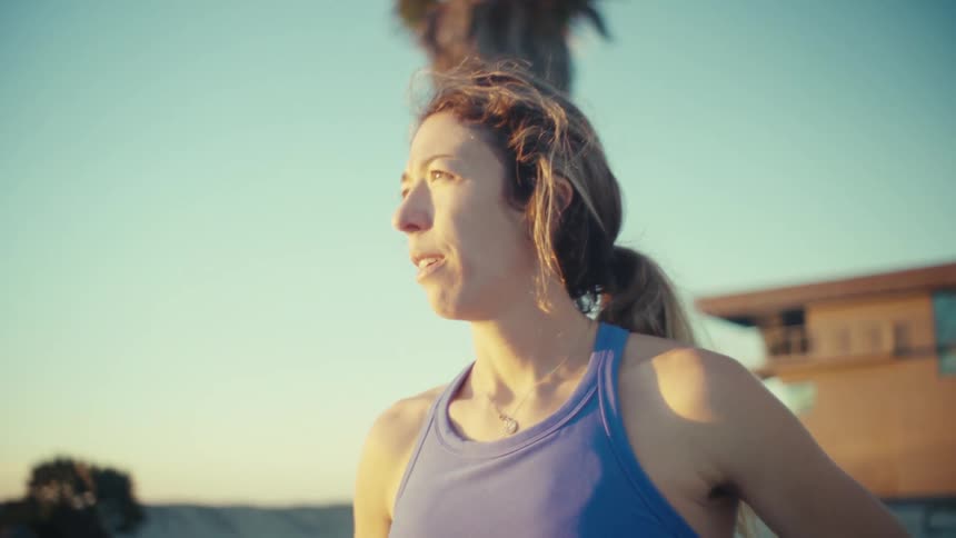 A pregnant woman in athletic wear stands on a beach at sunset catching her breath with her hands on her hips behind her palm trees and a lifeguard station are bathed in the warm golden light of the late afternoon