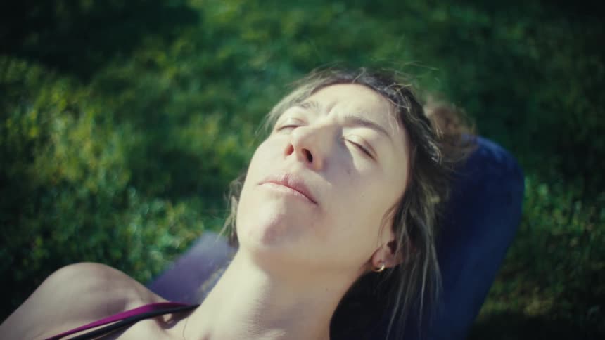A woman lies on a yoga mat in a sunlit grassy field with her eyes closed the camera slowly pulls back and rotates to a topdown view as she rests in the sun