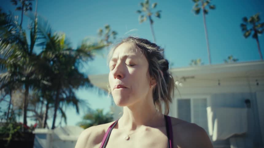 A woman in athletic wear drinks water from a clear bottle while standing outdoors in the sunlight after finishing she closes the bottle and leans down toward a table or bench