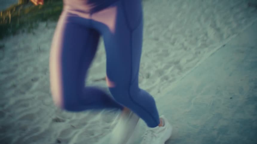 A woman wearing a blue sports top and leggings runs along a sandy beach path during a bright sunset while a building and palm trees stand in the background