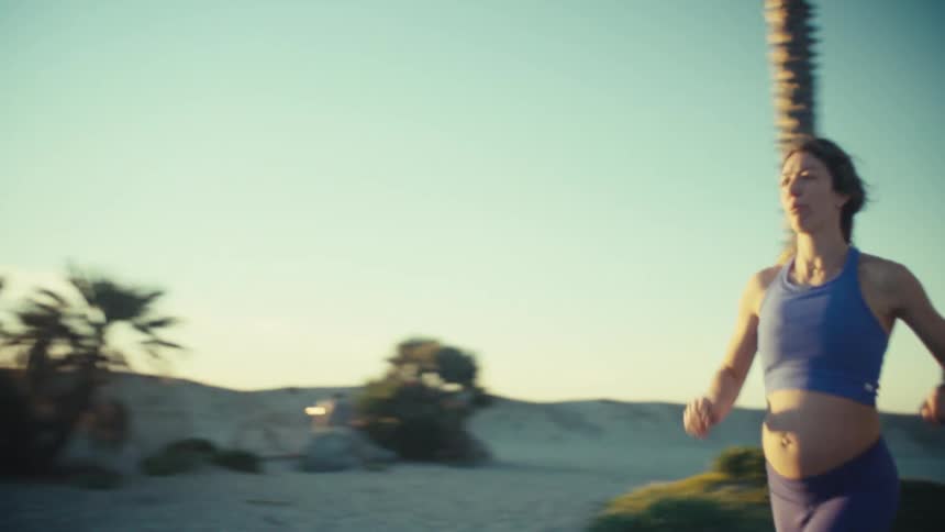 A woman wearing blue athletic clothing runs along a paved path at a beach during sunset with a lifeguard station and palm trees in the background