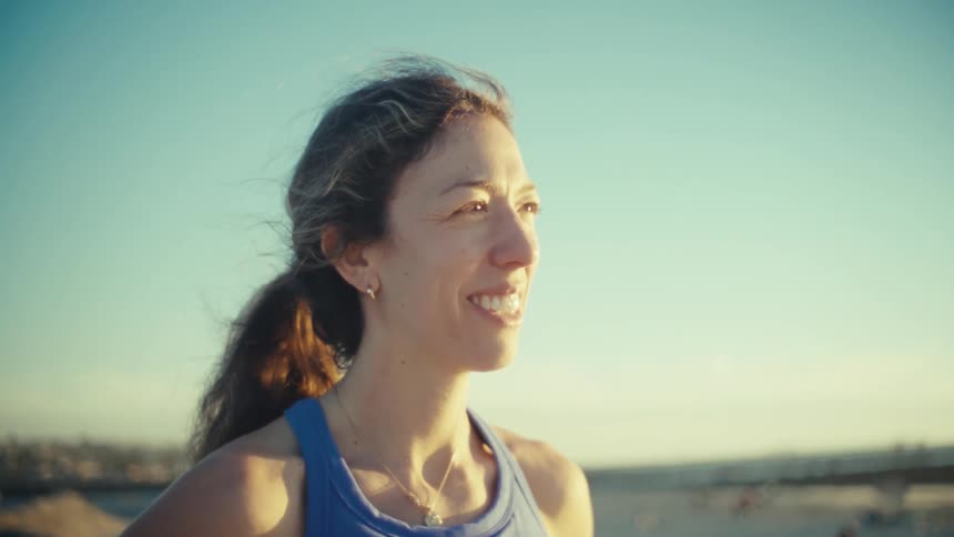 Woman jogging on the beach stops to rest looking around smiling as sunlight shines on her face