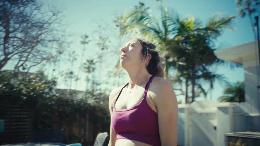 A woman wearing a purple sports bra and black leggings performs yoga on a mat in a sunny backyard by stretching her arms toward the sky while surrounded by green grass and palm trees