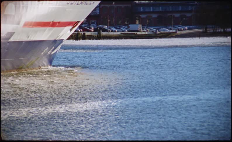 A large ships bow with painted lettering and a red stripe slices through calm blue water near a shoreline of parked cars and buildings leaving a foamy wake