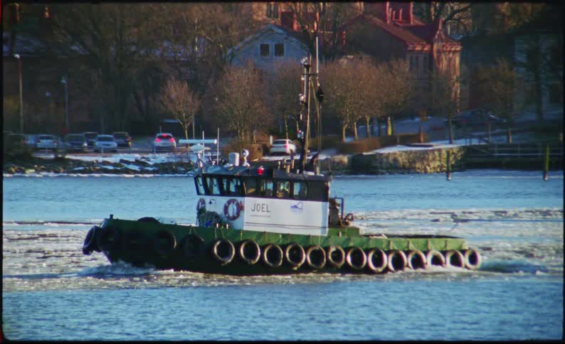 A green and white tugboat named joel moves across a cold icy body of water as the tugboat travels from left to right it reveals a large green barge being towed behind it