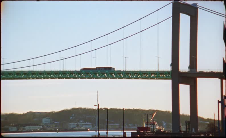 A cargo truck and other cars drive across a large green suspension bridge spanning over a wide river while distant hills and buildings sit under a clear sky