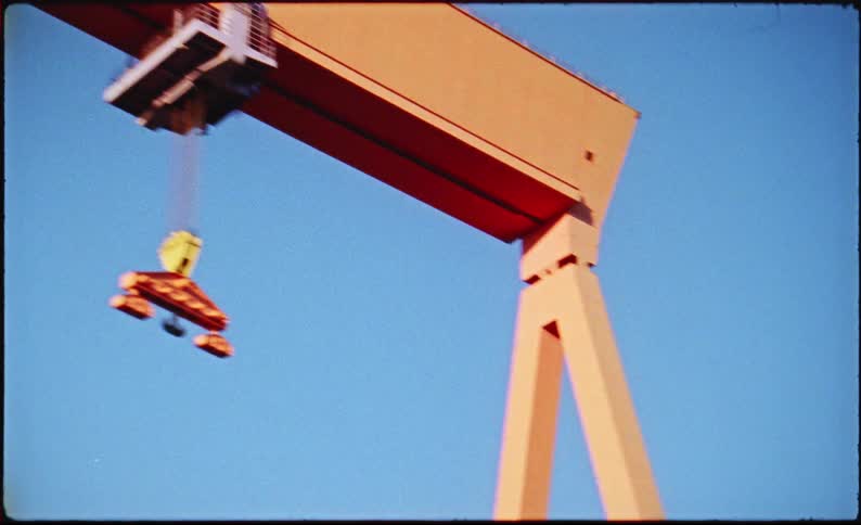A large orange gantry crane with the word eriksberg printed on it stands against a clear blue sky the cranes trolley moves along the horizontal beam while carrying a heavy lifting attachment suspended by cables