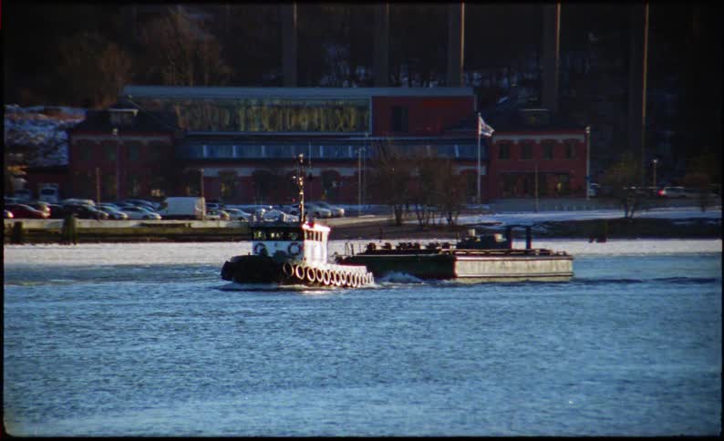 A seagull flies over a cold body of water followed by a tugboat pulling a barge past a distant city and suspension bridge