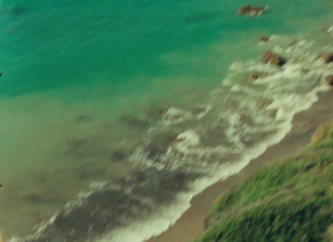 An elevated view shows turquoise ocean waves crashing against a sandy beach and a large rock formation