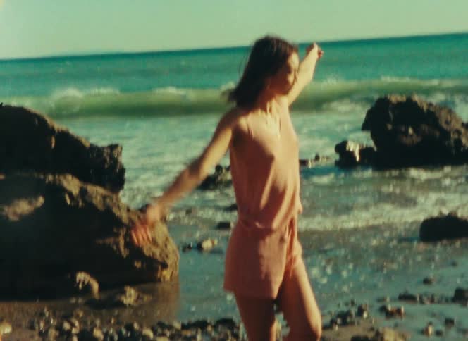 A woman wearing a peach colored tank top and matching shorts balances on one leg while standing on a rocky beach with blue ocean waves in the background