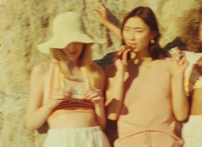Three young women relax against a sunlit rock wall while applying lip balm and smiling