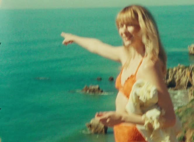 Teenager girl with blonde hair wearing an orange bikini walking on a cliff next to the ocean and pointing at the horizon while smiling outdoors during day time
