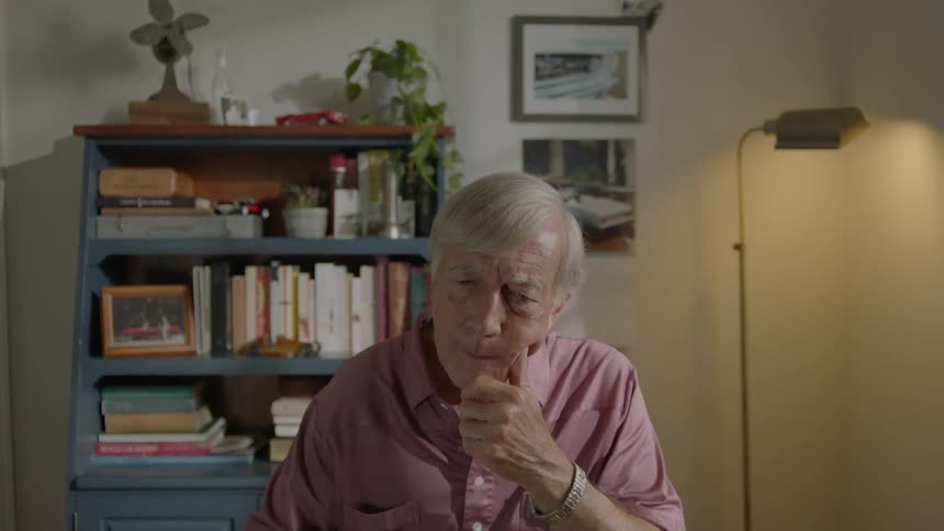 An elderly man sits in front of a lamp and bookshelf