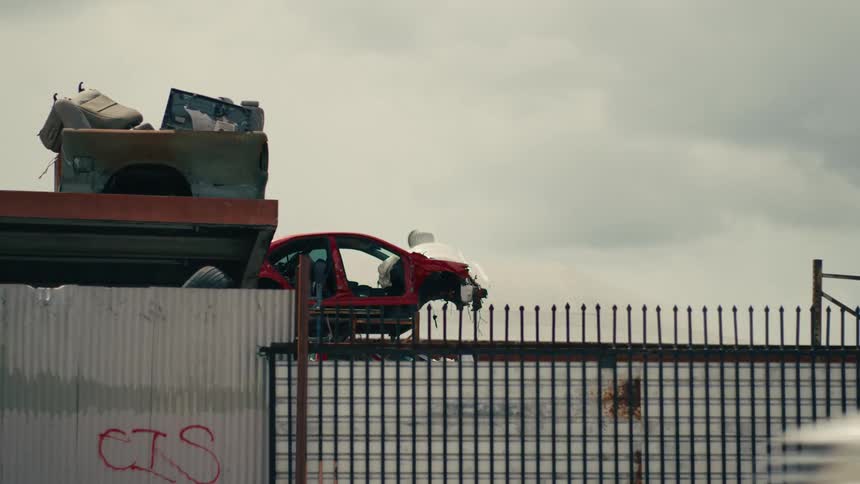 Red car shell on platform behind metal fence with white van passing in the foreground