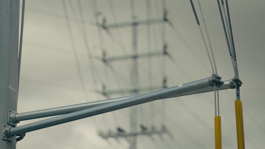 A utility pole features metal support arms and heavy electrical cabling against a backdrop of a large transmission tower under an overcast sky