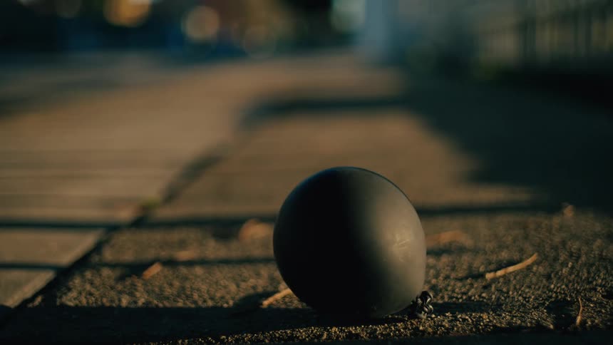 A single black balloon rests on a concrete surface as long dark shadows from a fence stretch across the ground in a dim outdoor setting