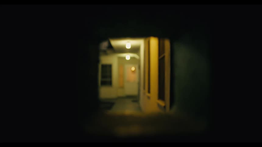 A view through a small square opening in a dark wall reveals a blurry outdoor hallway with yellow walls and bright lights at night