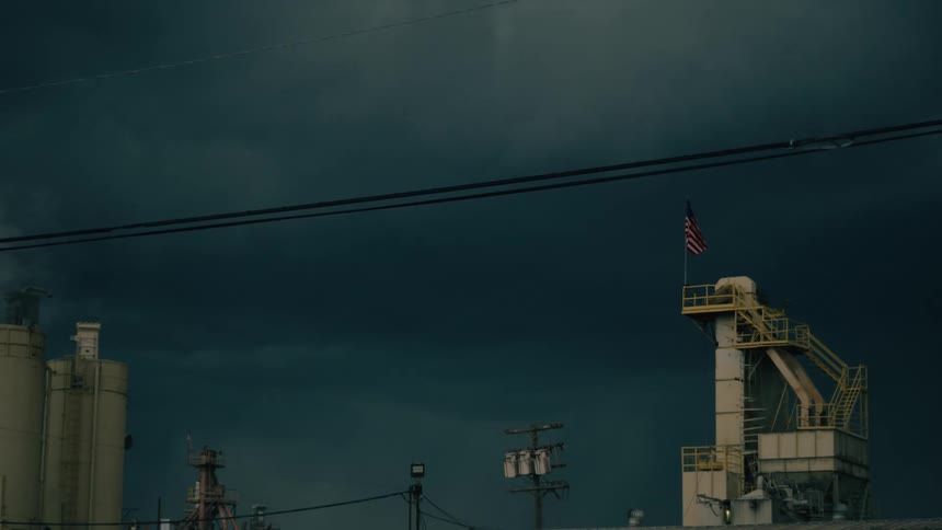 Industrial tower with a staircase and flag hoisted under a dark sky with electrical wires running through the panel