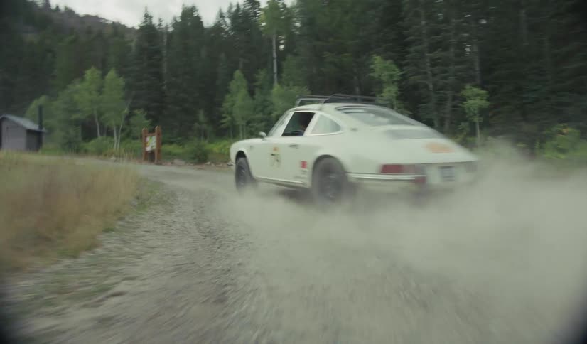 A car speeds along a gravel road through a forested clearing throwing up a thick cloud of dust that nearly obscures a small cabin and signpost in the background