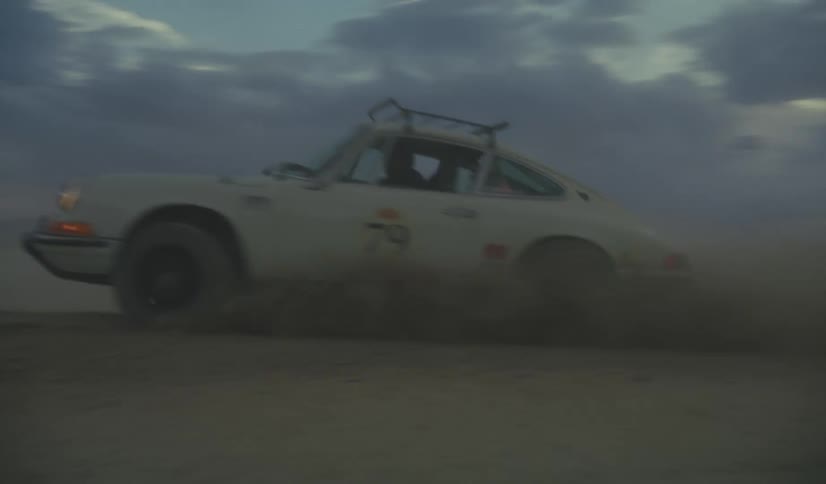 A white vintage sports car with a roof rack drives fast across a flat desert landscape at dusk kicking up a large cloud of dust under a cloudy purple and blue sky