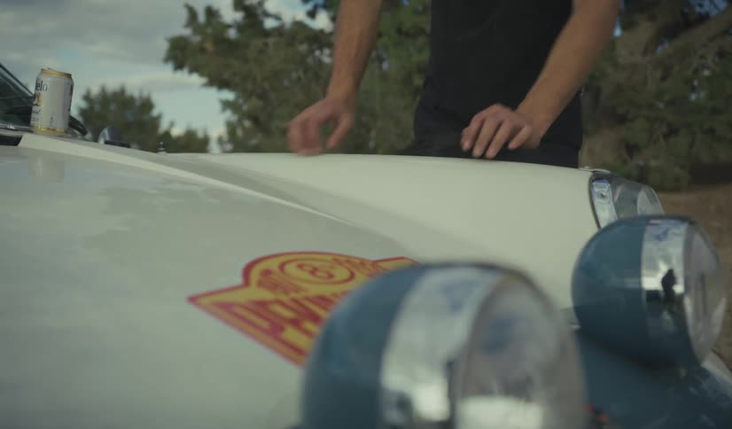A man wearing a black shirt leans his hands against the hood of a vintage white car parked outdoors
