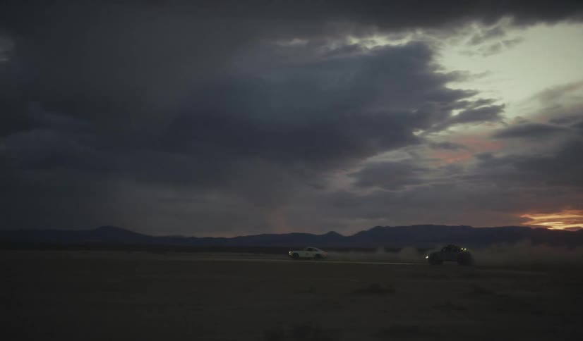Vehicles with headlights on cross a desert plain under storm clouds at dusk