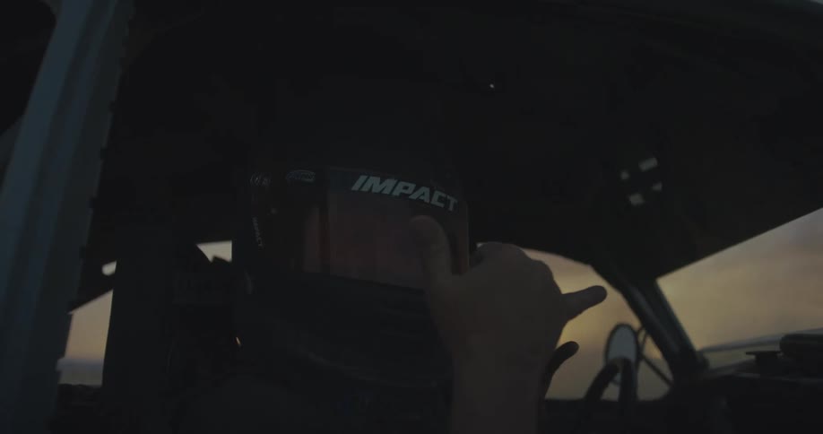 A person wearing a black racing helmet and suit sits inside a vehicle and makes a hand gesture while looking toward the camera during dusk